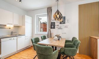 A modern kitchen with a dining table and green chairs. The walls are light and there is plenty of natural light coming through the window.