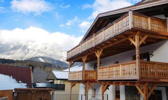 A charming wooden house with several balconies and a beautiful view of the mountains. In the foreground lies the snow-covered ground.