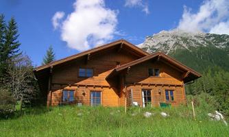 A charming wooden house surrounded by green meadows and trees. In the background, majestic mountains and a clear sky can be seen.