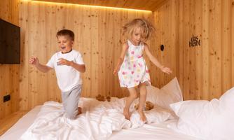 Two children are happily jumping on a bed in a cozy wooden room. The walls are made of wood, and there is a warm, inviting atmosphere.