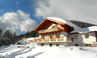 A beautifully located house in the snow with a wooden balcony. The surroundings are filled with trees and a clear blue sky.