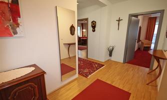 A bright hallway with wooden flooring and a red carpet. A mirror and a piece of art hang on the wall, while a cabinet and a plant stand in the room.