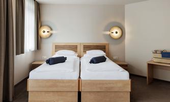 A modern bedroom with a bed. The walls are light, and there are large windows with curtains and simple wooden furniture.