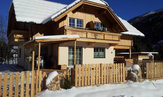 A beautiful wooden house in winter, surrounded by snow. The sky is clear and blue, and a fence surrounds the property.
