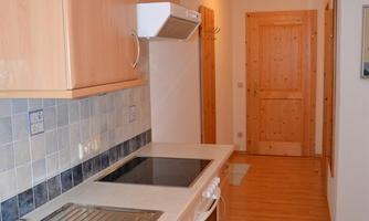 A modern kitchen with light wooden furniture and a clear work surface. The room transitions into a long hallway that leads to the entrance door.