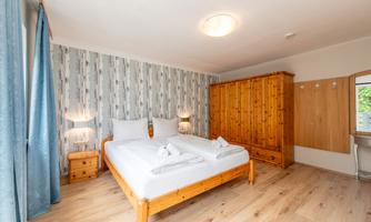 A cozy bedroom with a double bed and wooden furniture. The walls are decorated with a modern pattern, and the room has large windows with blue curtains.