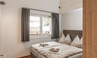 A cozy bedroom with a double bed and wooden flooring. The windows are dressed with curtains and provide light in the room.