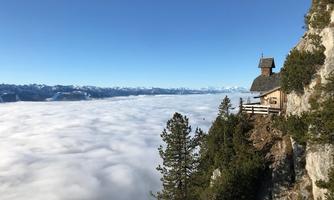 A picturesque mountain panorama with a small cabin on a steep slope. In front of the cabin, a dense blanket of clouds spreads out, framing the peaks of the mountains.
