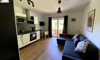 A modern living kitchen with a cozy sofa and a dining area. Large windows let in plenty of light and provide a view outside.