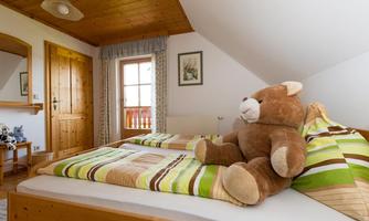 A cozy bedroom with two beds and a large plush bear. The room has wooden ceilings and a window with a view outside.