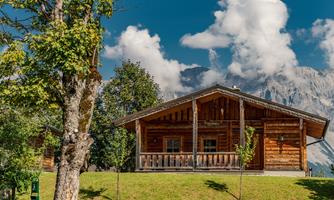 A cozy wooden house surrounded by trees and green spaces. In the background, majestic mountains and a blue sky with white clouds can be seen.