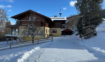 A cozy house in the snow, surrounded by trees. The sky is clear and blue, creating a pleasant winter atmosphere.
