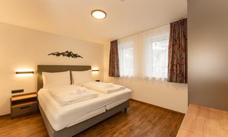 A modern bedroom with a double bed and wooden floors. Large windows with curtains provide natural light.