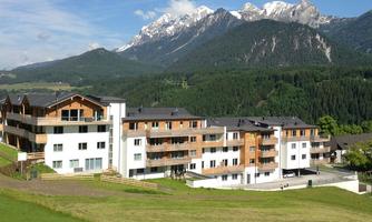 A modern residential complex with a wooden and concrete structure, surrounded by green meadows. Impressive mountains can be seen in the background.
