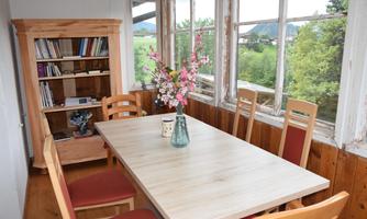 A bright dining room with a large table and chairs. On the table sits a vase with flowers and in the corner there is a bookshelf.
