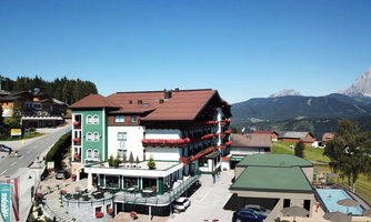 A hotel building with a red roof and several balconies. In the background, green mountains and a clear blue sky can be seen.