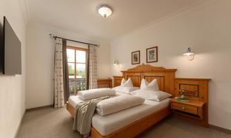 A cozy bedroom with a large double bed and warm wooden furniture. The room is illuminated by a ceiling lamp and two wall lamps.