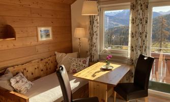 A cozy room with wooden paneling and a window view of the mountains. A table with two chairs and a small flower creates an inviting atmosphere.