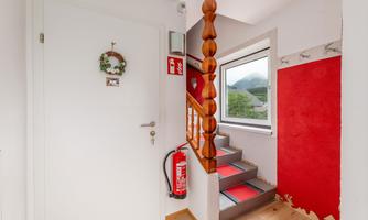 A bright hallway with a wooden staircase and a red wall area. A fire extinguisher and a small decoration hang at the entrance.