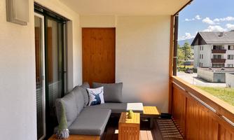 A cozy balcony with a comfortable seating area and wooden floor. In the background, houses and a clear blue sky can be seen.