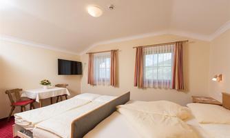 A cozy bedroom with two beds and a table. The room is bright with windows and a TV on the wall.
