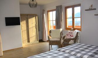A cozy room with a bed and a wicker chair.  
Large windows let in plenty of light and offer a view outside.