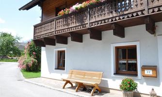 A traditional house with a wooden balcony decorated with flowers. In front of the house, there is a wooden bench next to the windows.