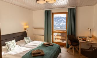 A cozy bedroom with two beds and a view of the mountains. The room is modernly furnished, with a desk area and large windows.