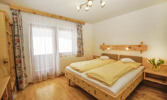 A cozy bedroom with a double bed and wooden furniture. Large windows let in plenty of daylight, and a fresh flower sits on the nightstand.
