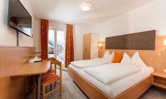 A bright bedroom with a double bed and orange accents. There is a wooden desk and a television on the wall.