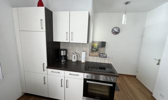A modern kitchen with white cabinets and a black countertop. It is well-equipped with a stove, coffee maker, and kettle.