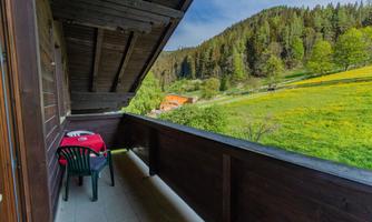 A balcony with a tablecloth and two chairs. In the background, green meadows and forests can be seen.