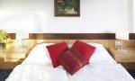 A cozy bedroom with a large bed and red pillows. A painting hangs on the wall, and the room is brightly lit.