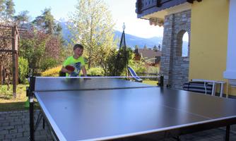 A boy is playing table tennis in the garden. In the background, there are trees and a beautiful mountain view.