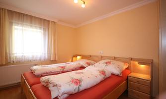 A cozy bedroom with two beds and floral bedcovers. The room has large windows and a warm, inviting atmosphere.