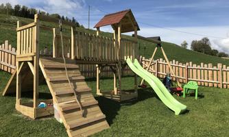 A wooden playground with a slide, a ladder, and a climbing frame. The playground is situated on a meadow in a green environment.