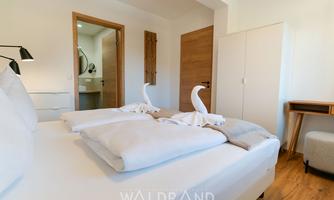 A modern bedroom with a double bed and decorative towels shaped like swans. The room features bright walls, a wooden floor, and natural light sources.