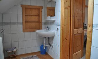 A small bathroom with white tiles and wooden details. There is a sink, a mirror, and a wooden shelf.