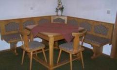 A cozy dining area with a round wooden table and wooden chairs. The bench is equipped with patterned cushions and the walls are plain.