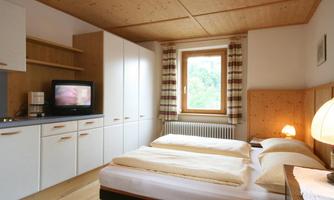 A simple bedroom with two beds, a television, and bright wood interiors. A window with curtains allows for natural light.