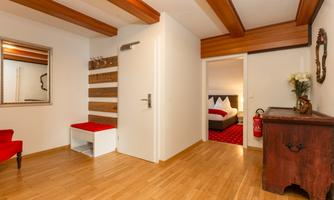 A modern entrance area with wooden flooring and bright walls. On the left side, there is a red bench and a mirror, while a bedroom is visible in the background.