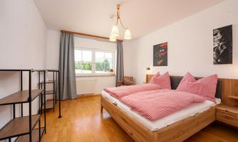 A modern bedroom with a comfortable double bed and red pillows. There are large windows that let in plenty of daylight, as well as a simple furniture arrangement.