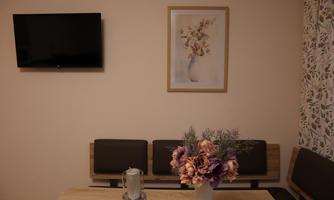 A cozy dining area with a wooden table and a vase full of flowers. A picture hangs on the wall and a television is mounted.