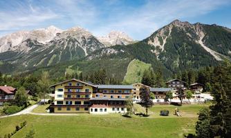 A picturesque mountain panorama with a large hotel building in the foreground. The surroundings are green with trees and a clear blue sky color.