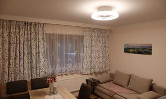 A cozy living room with a beige sofa and a dining table. The window is decorated with blooming curtains and a picture of a landscape hangs on the wall.