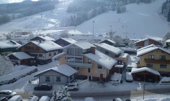 A snowy landscape with a residential area and surrounding mountains. The houses are covered in snow, and the sun is shining through the clouds.
