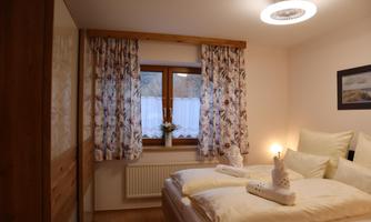 A cozy bedroom with a double bed and bright bedcovers. Large windows with floral curtains provide plenty of daylight.