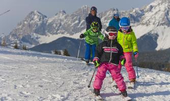 A group of children is skiing on a snow-covered slope. Impressive mountains are visible in the background.