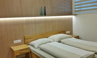 A modern bedroom with a wooden bed and two mattresses. The walls are light and equipped with indirect lighting.