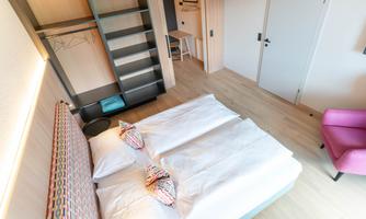 A modern hotel room with a double bed, bright walls, and a pink armchair. The decor is light and inviting, perfect for a relaxing stay.